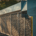 A modern security and privacy barrier system photographed in warm evening light. The fence features modular panels filled with stacked gray stone (gabion-style), framed by clean silver metal posts and horizontal caps. A solid metal panel section appears on the right, accented by a yellow vertical post. Barbed wire runs along the top. The barrier sits beside a gravel path with soft dust in the air and green foliage visible on the left.