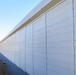 A long, modern white security and privacy barrier wall with horizontal paneling stretches into the distance along a gravel path. The clean, solid panels are framed with vertical metal supports and topped by a continuous cap. Overhead power lines are visible against a clear blue sky, with sparse vegetation growing at the base of the wall.